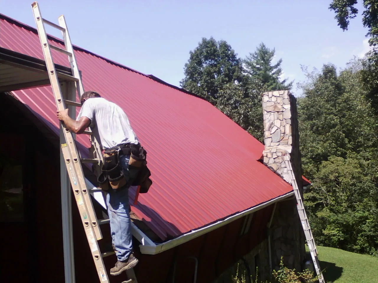 Roofer on a ladder beside a red metal roof and stone chimney — Soffit & Fascia Repair by Roofing Brothers in Buckhannon